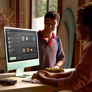 พนักงานคนหนึ่งนั่งอยู่หลังเคาน์เตอร์ในร้านขายกระดานโต้คลื่นและสินค้าอื่นๆ กำลังใช้ iMac เพื่อดำเนินการชำระเงินให้กับลูกค้าที่อยู่ฝั่งตรงข้ามเคาน์เตอร์
