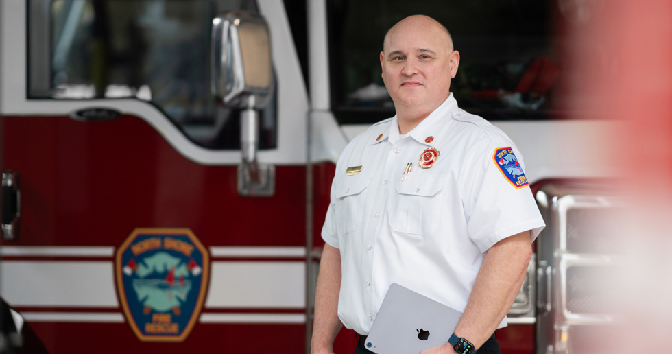 The fire chief stands in front of a fire truck holding an iPad and wearing an Apple Watch